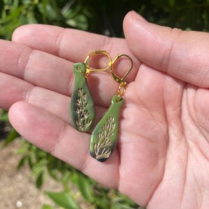 May include: A pair of green ceramic earrings with gold leaf detailing. The earrings are shaped like teardrops and feature a delicate floral design.