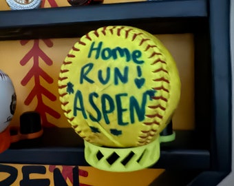 May include: Yellow softball with black stitching and the words "Home Run! Aspen" written in green. The softball is displayed in a black baseball display case with red stitching.