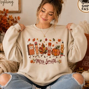 May include: A beige crewneck sweatshirt featuring a graphic print of a smiling notebook, pencil, maple leaf, glue stick, pumpkin, and another pencil. The text Mrs. Smith is printed in a dark red script font below the illustrated characters.
