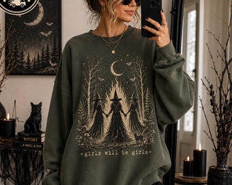 May include: An olive green crewneck sweatshirt featuring a graphic print of three witches holding hands in a forest under a crescent moon. The design includes trees, stars, and the text "girls will be girls" in a light beige color.