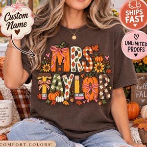 May include: A dark brown short-sleeve t-shirt featuring a graphic print with the text MRS TAYLOR. The design includes orange pumpkins, pencils, autumn leaves, and flowers in various patterns like plaid and floral, accented with pink bows.
