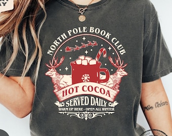 May include: A charcoal gray t-shirt featuring a graphic print with the text North Pole Book Club, Hot Cocoa Served Daily, and Warm Up Here - Open All Winter. The design includes a red mug with marshmallows, a candy cane, and two reindeer.