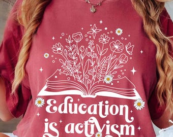 Puede incluir: Una camiseta color granate con un gráfico blanco de un libro abierto con flores que crecen de él. El texto "Education is activism" está escrito debajo del libro.
