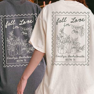May include: A charcoal gray t-shirt and a cream t-shirt, each featuring a back graphic of a postage stamp outline containing the text fall Love in, Emily's Bachelorette, and AUSTIN, TX, with illustrations of pumpkins, acorns, and autumn leaves.