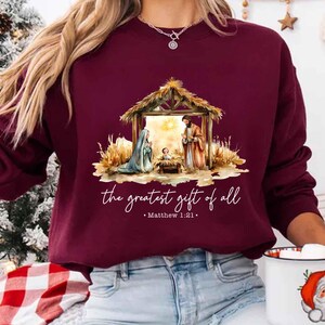 May include: Maroon sweatshirt featuring a Nativity scene with the text "the greatest gift of all" and Matthew 1:21. The design includes Mary, Joseph, and baby Jesus in a manger. The sweatshirt is a long-sleeved crew neck.