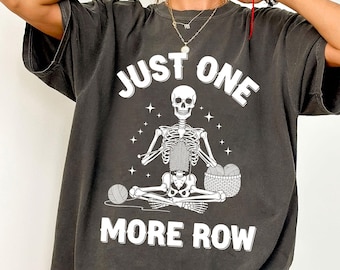 May include: Dark gray t-shirt featuring a white skeleton knitting with the text "JUST ONE MORE ROW". The skeleton is seated in a cross-legged position, holding a basket of yarn and a ball of yarn. The shirt has a relaxed fit.