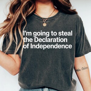 May include: Dark gray t-shirt with the text "I'm going to steal the Declaration of Independence" in white. The shirt is a crew neck and has short sleeves. The person is wearing blue jeans.