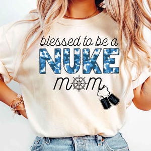 May include: Cream-colored t-shirt with the text "blessed to be a NUKE mom" in black and blue. The word "NUKE" is in blue camouflage print. Includes a ship's wheel graphic and dog tags.