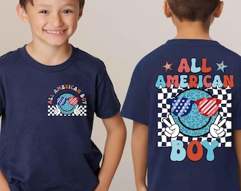 May include: Navy blue t-shirt with a graphic design featuring a smiley face wearing sunglasses in red, white, and blue. The shirt has the text "ALL AMERICAN BOY" and a peace sign. The shirt is for children.
