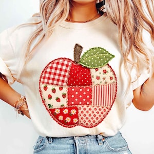 May include: A cream-colored short-sleeve t-shirt featuring a large, central apple-shaped graphic. The apple is constructed from a patchwork of various red and white fabric patterns, including gingham, polka dots, strawberries, and daisies.