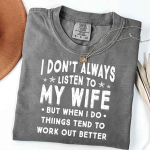 May include: A gray Comfort Colors t-shirt with the text "I DON'T ALWAYS LISTEN TO MY WIFE BUT WHEN I DO THINGS TEND TO WORK OUT BETTER" in white. A silver necklace with a pendant and a brown hat are also visible.