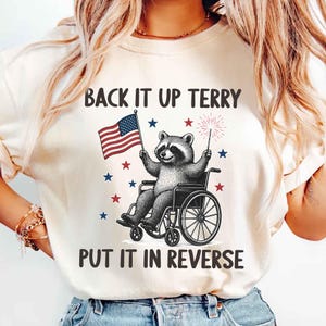 May include: Cream-colored t-shirt featuring a raccoon in a wheelchair holding an American flag and a sparkler. The text "BACK IT UP TERRY PUT IT IN REVERSE" is printed above and below the raccoon illustration, with red and blue stars.