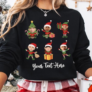May include: Black sweatshirt featuring a festive design of elf characters with personalized faces, wearing red and green outfits and Santa hats. The words "Your Text Here" are printed in white script at the bottom of the sweatshirt. A Christmas-themed apparel item.