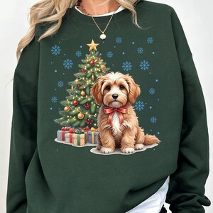May include: Dark green sweatshirt featuring a Christmas tree, presents, and a cute dog wearing a red bow. The tree is decorated with ornaments and lights, and snowflakes are scattered around the design. A festive holiday-themed sweatshirt.