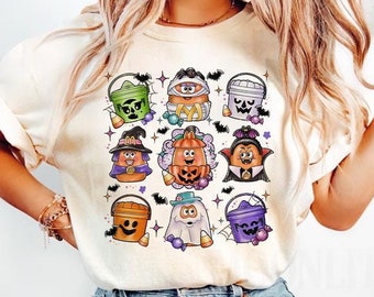 May include: A cream-colored t-shirt featuring a graphic print of nine cartoon characters dressed in Halloween costumes, including various buckets, a mummy, a witch, a pumpkin, a vampire, and a ghost, surrounded by small bats and candy corn.