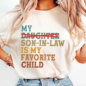 May include: Cream-colored t-shirt with the text "My Son-in-Law is My Favorite Child" in various colors. The word "Daughter" is crossed out with a red line. The shirt has a relaxed fit.