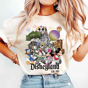 May include: A cream-colored t-shirt featuring a graphic print of Disney characters playing musical instruments in front of a castle, a geodesic sphere, and a train. The text reads Disneyland Est. 1955, accompanied by fireworks and a hot air balloon.