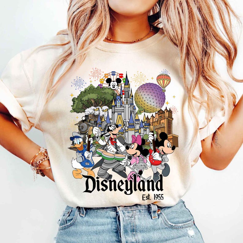 May include: A cream-colored t-shirt featuring a graphic print of Disney characters playing musical instruments in front of a castle, a geodesic sphere, and a train. The text reads Disneyland Est. 1955, accompanied by fireworks and a hot air balloon.