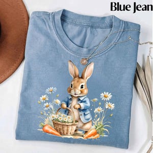 May include: A blue jean t-shirt with a cute cartoon bunny rabbit holding a basket of daisies. The bunny is wearing a blue jacket and has brown fur. The t-shirt has a distressed look.