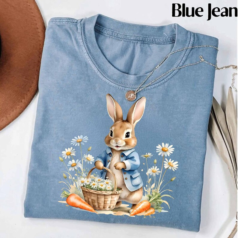 May include: A blue jean t-shirt with a cute cartoon bunny rabbit holding a basket of daisies. The bunny is wearing a blue jacket and has brown fur. The t-shirt has a distressed look.