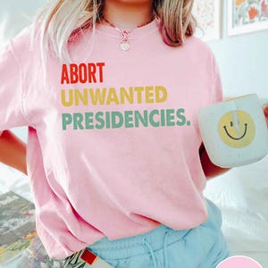 May include: A light pink t-shirt with the words "ABORT UNWANTED PRESIDENCIES" in red, yellow, and green. A white mug with a yellow smiley face is held in the right hand.
