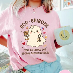 May include: Pink t-shirt with a cartoon ghost holding a prescription bottle and a stethoscope. Text on the shirt reads "BOO-SPIRONE" and "TAKE AS NEEDED FOR SPOOKY SEASON ANXIETY." A white mug with a yellow smiley face is also visible.
