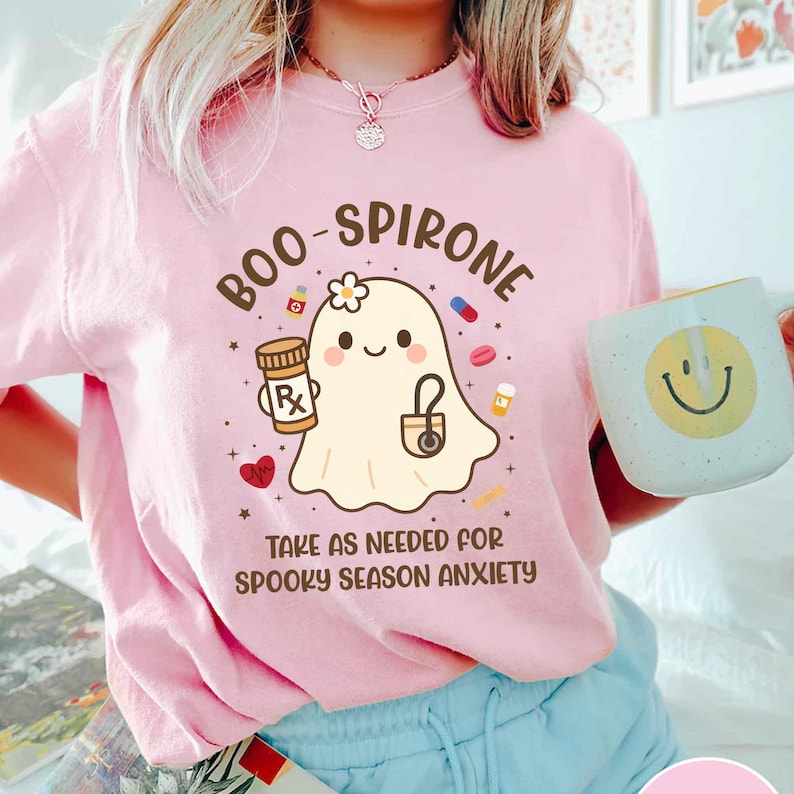 May include: Pink t-shirt with a cartoon ghost holding a prescription bottle and a stethoscope. Text on the shirt reads "BOO-SPIRONE" and "TAKE AS NEEDED FOR SPOOKY SEASON ANXIETY." A white mug with a yellow smiley face is also visible.