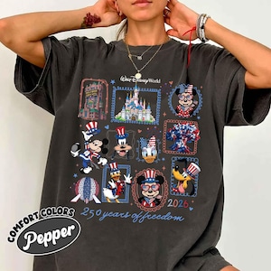 May include: A charcoal grey t-shirt featuring a graphic print with the text Walt Disney World and 250 years of freedom. The design includes illustrations of a castle, a hotel, and various cartoon characters wearing patriotic red, white, and blue hats.
