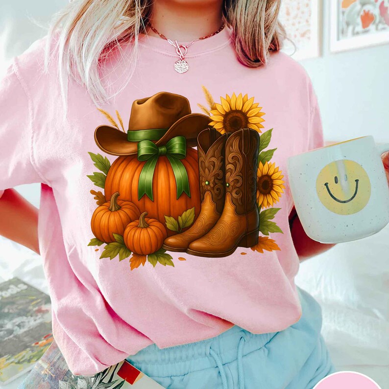 May include: Pink t-shirt featuring a fall-themed graphic. The design includes a brown cowboy hat with a green ribbon, an orange pumpkin, brown cowboy boots, sunflowers, and small pumpkins. A white mug with a smiley face is also visible.