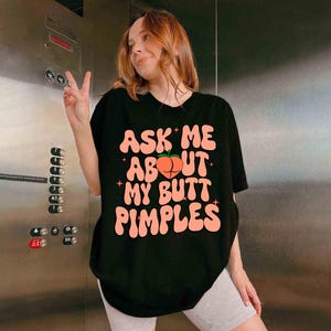 Op de afbeelding: Zwart t-shirt met de tekst "ASK ME ABOUT MY BUTT PIMPLES" in roze, met een perzikafbeelding. Het t-shirt wordt door een persoon gedragen.