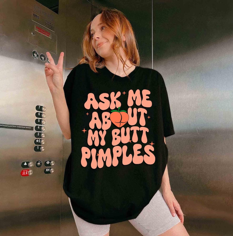 Op de afbeelding: Zwart t-shirt met de tekst "ASK ME ABOUT MY BUTT PIMPLES" in roze, met een perzikafbeelding. Het t-shirt wordt door een persoon gedragen.