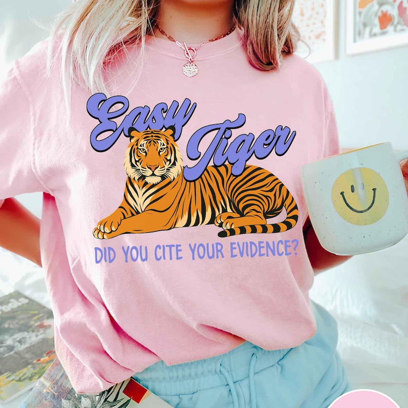 May include: A light pink t-shirt with the words "Easy Tiger" in a purple font, and a graphic of a tiger. The phrase "Did you cite your evidence?" is printed below the tiger. A white mug with a smiley face is visible.