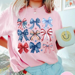 May include: A pink t-shirt with a design of red, white, and blue bows with stars. The bows are arranged in a pattern on the front of the shirt.