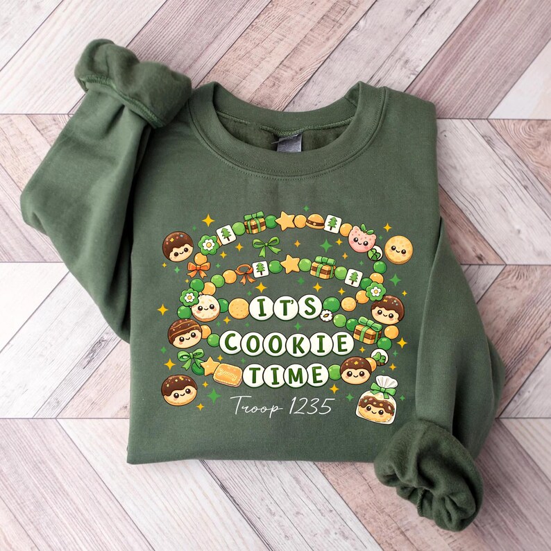 May include: A green sweatshirt with the text "It's Cookie Time" in white, arched above "Troop 1235". The design includes cartoon cookies, gifts, and stars. The sweatshirt is folded on a wooden surface.