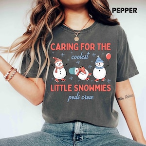 May include: Dark gray t-shirt with the text "CARING FOR THE coolest LITTLE SNOWMIES peds crew." The shirt features three cartoon snowmen wearing hats and scarves, ice skating, and holding a balloon. The word "PEPPER" is at the top right.