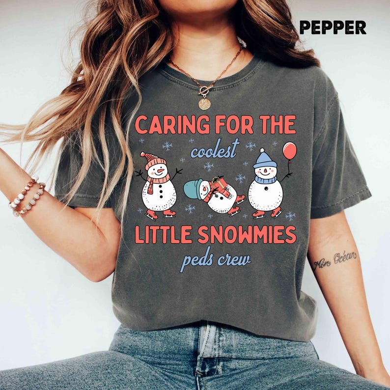 May include: Dark gray t-shirt with the text "CARING FOR THE coolest LITTLE SNOWMIES peds crew." The shirt features three cartoon snowmen wearing hats and scarves, ice skating, and holding a balloon. The word "PEPPER" is at the top right.