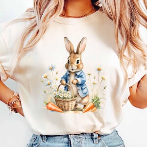 May include: A white t-shirt with a cartoon illustration of a brown bunny rabbit wearing a blue jacket and holding a basket of white flowers. The bunny is standing in a field of white flowers and green grass. There are two orange carrots on the ground.