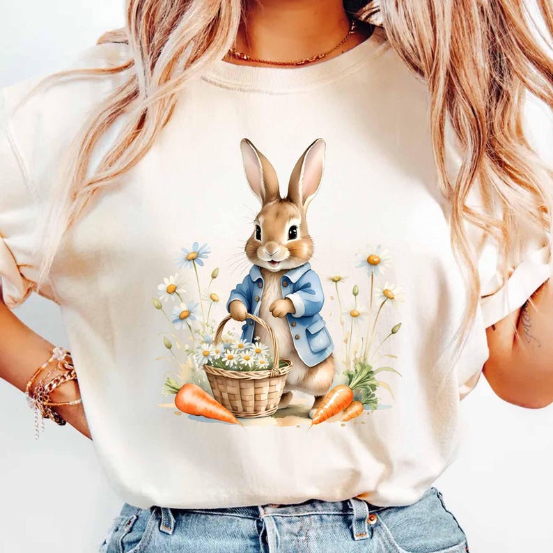 May include: A white t-shirt with a cartoon illustration of a brown bunny rabbit wearing a blue jacket and holding a basket of white flowers. The bunny is standing in a field of white flowers and green grass. There are two orange carrots on the ground.