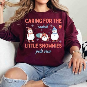 May include: Burgundy sweatshirt with the text "Caring for the coolest Little Snowmies peds crew." The graphic features three snowmen wearing hats and scarves, ice skating, and holding a balloon. The sweatshirt is a long-sleeved crew neck.