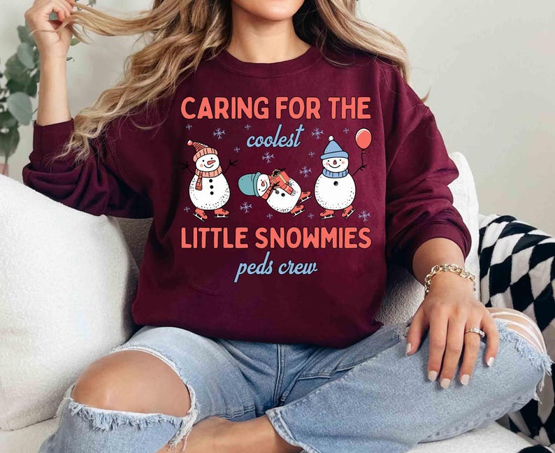 May include: Burgundy sweatshirt with the text "Caring for the coolest Little Snowmies peds crew." The graphic features three snowmen wearing hats and scarves, ice skating, and holding a balloon. The sweatshirt is a long-sleeved crew neck.
