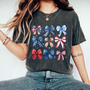 May include: A dark gray t-shirt with a pattern of red, white, and blue bows. The bows are arranged in a grid pattern and have a watercolor effect. The shirt has a relaxed fit and a crew neckline.