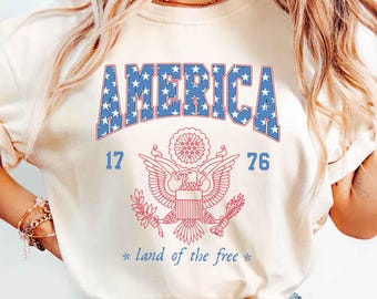 May include: A white t-shirt with a blue and red graphic print of the word "AMERICA" with stars, the American eagle, and the dates "17" and "76". The text "land of the free" is printed below the graphic.