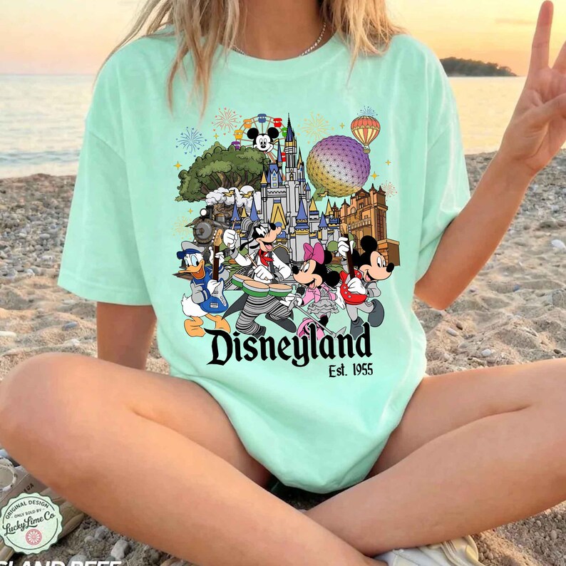 May include: A light mint green t-shirt featuring a colorful graphic of Disney characters playing instruments in front of a castle, with the text Disneyland Est. 1955. The design includes Mickey Mouse, Minnie Mouse, Donald Duck, and Goofy.