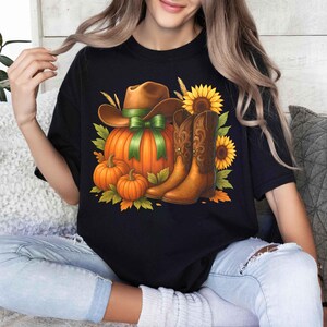 May include: Black t-shirt featuring a fall-themed graphic. The design includes a brown cowboy hat with a green ribbon, an orange pumpkin, brown cowboy boots, sunflowers, and smaller pumpkins. The artwork has a detailed, illustrative style.