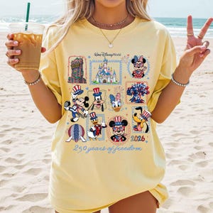 May include: A pale yellow t-shirt featuring a graphic print with Walt Disney World text, a castle, and various cartoon characters wearing patriotic red, white, and blue outfits. The design includes the text 250 years of freedom and 2026.