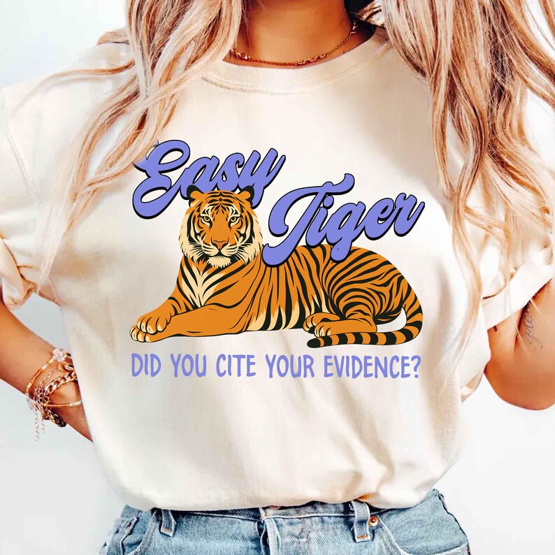 May include: Cream-colored t-shirt featuring a tiger illustration with orange and black stripes. The text "Easy Tiger" is above the tiger, and below it reads "DID YOU CITE YOUR EVIDENCE?" in a purple font. The shirt has short sleeves.