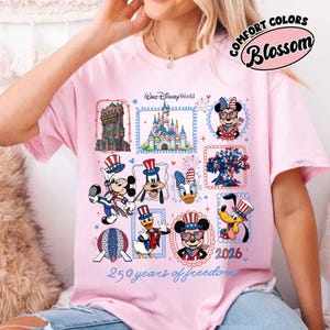 May include: A light pink t-shirt featuring a graphic print of Walt Disney World characters and landmarks with patriotic red, white, and blue accents. The design includes the text Walt Disney World, 2026, and 250 years of freedom.