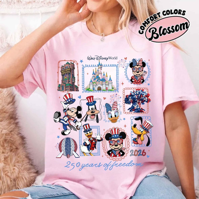 May include: A light pink t-shirt featuring a graphic print of Walt Disney World characters and landmarks with patriotic red, white, and blue accents. The design includes the text Walt Disney World, 2026, and 250 years of freedom.
