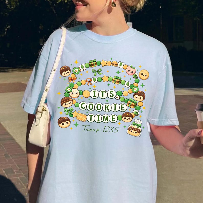 May include: Light blue t-shirt with a colorful graphic that says "Its Cookie Time" in a rainbow design. The design includes cartoon cookies, stars, and gift boxes. The text "Troop 1235" is below the main text.