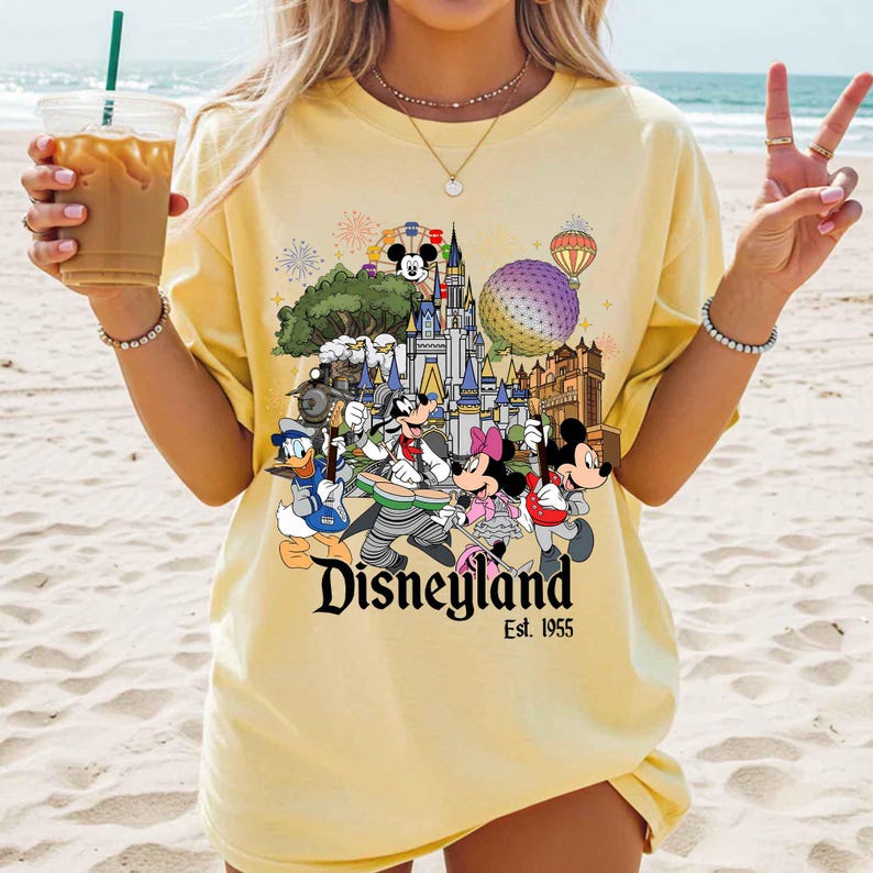 May include: A pale yellow t-shirt featuring a colorful graphic of Disney characters playing musical instruments in front of a castle. The print includes Mickey Mouse, Minnie Mouse, Donald Duck, and Goofy, with the text Disneyland Est. 1955.
